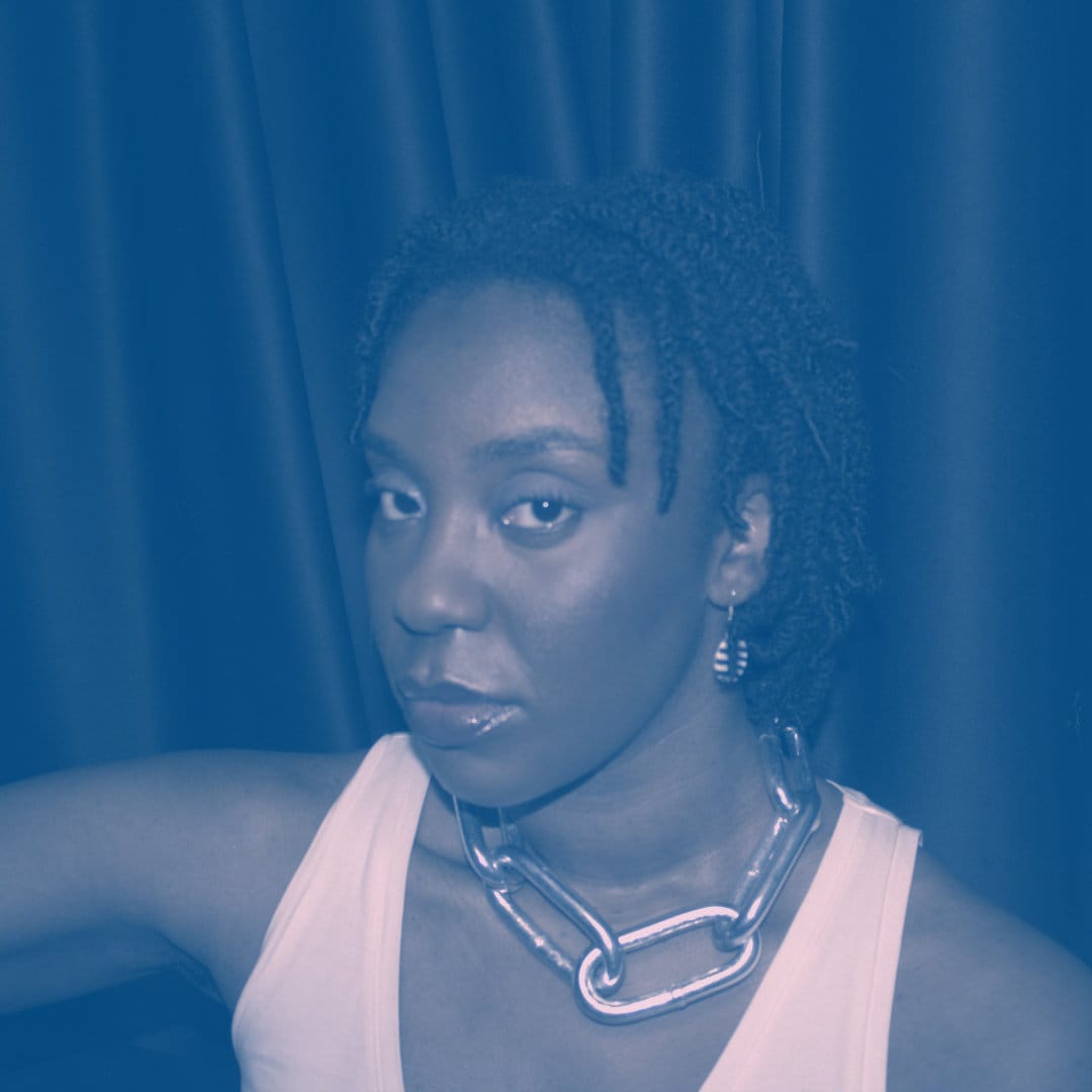 A black woman with twists, wearing a white vest and silver chain against black curtains.