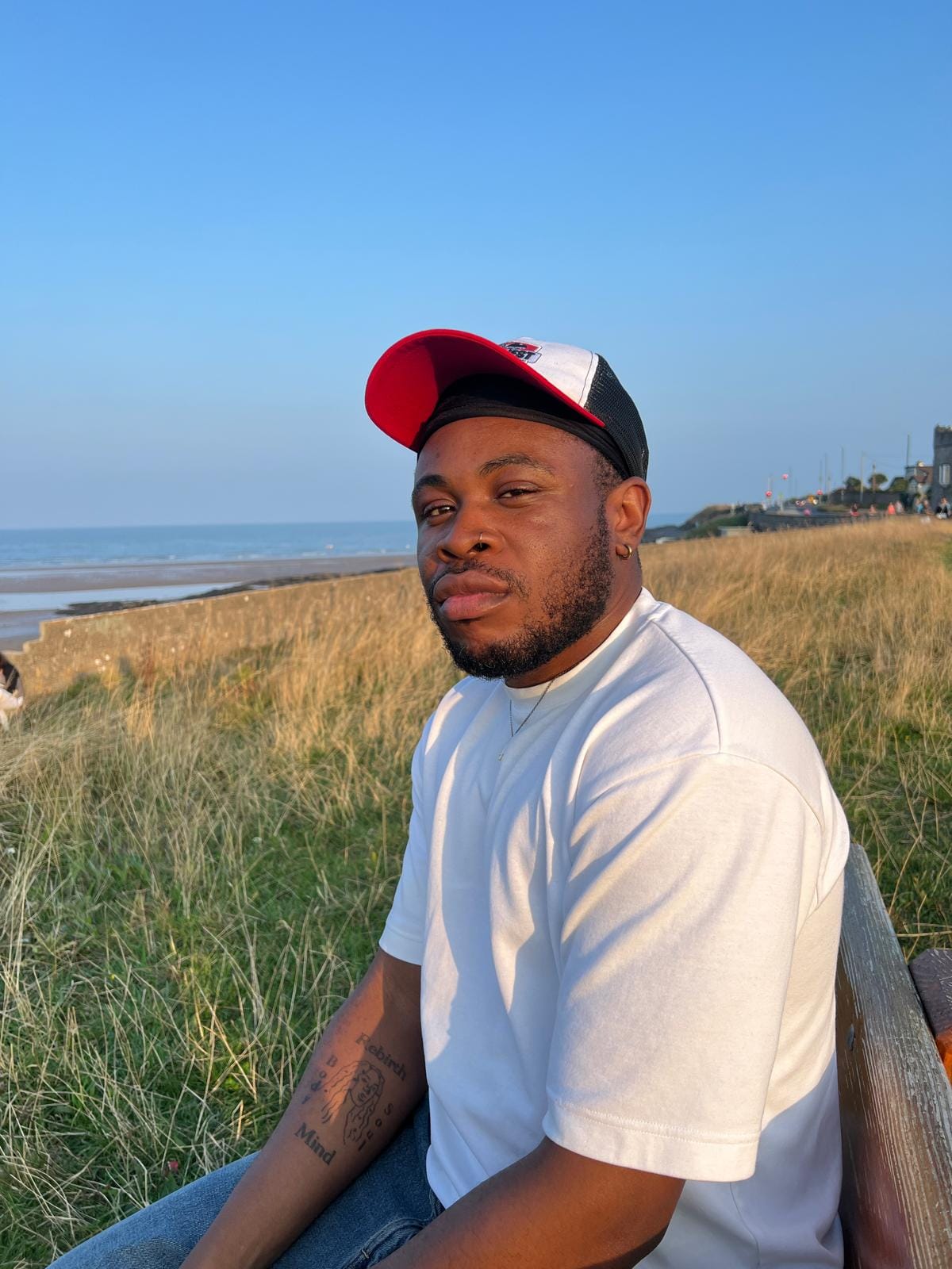 A black man with a nose ring, wearing a white shirt and red/white/black cap sitting on a bench outside in a grassy area.