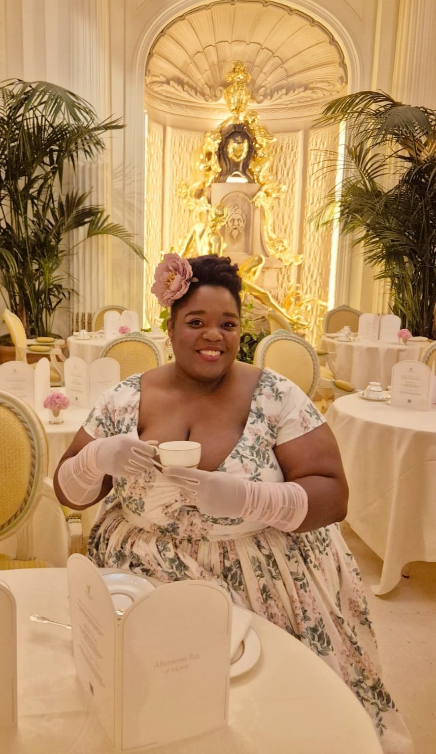 A fat black femme in a restaurant holding a teacup, wearing a flowery dress and pink gloves.