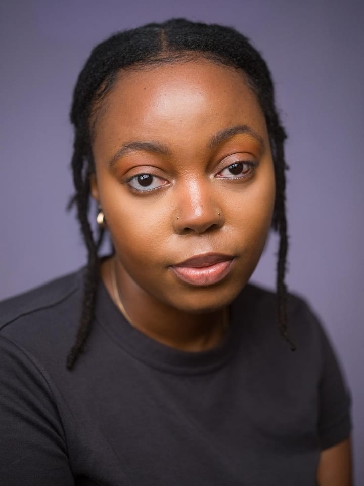 A South African person with brown eyes looking into the camera, she is wearing a black top with gold jewellery. 